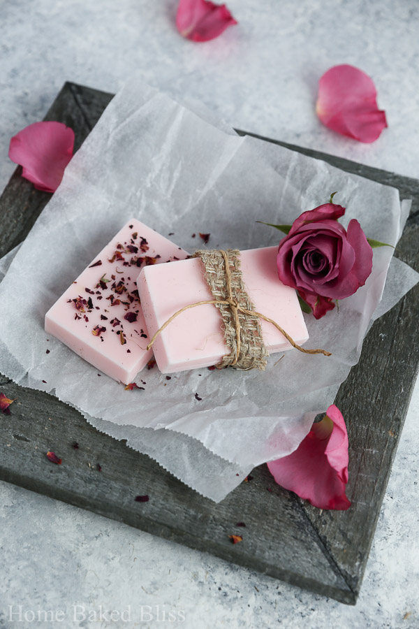 Two bars of diy rose soap on a wooden plate decorated with twine and rose petals.